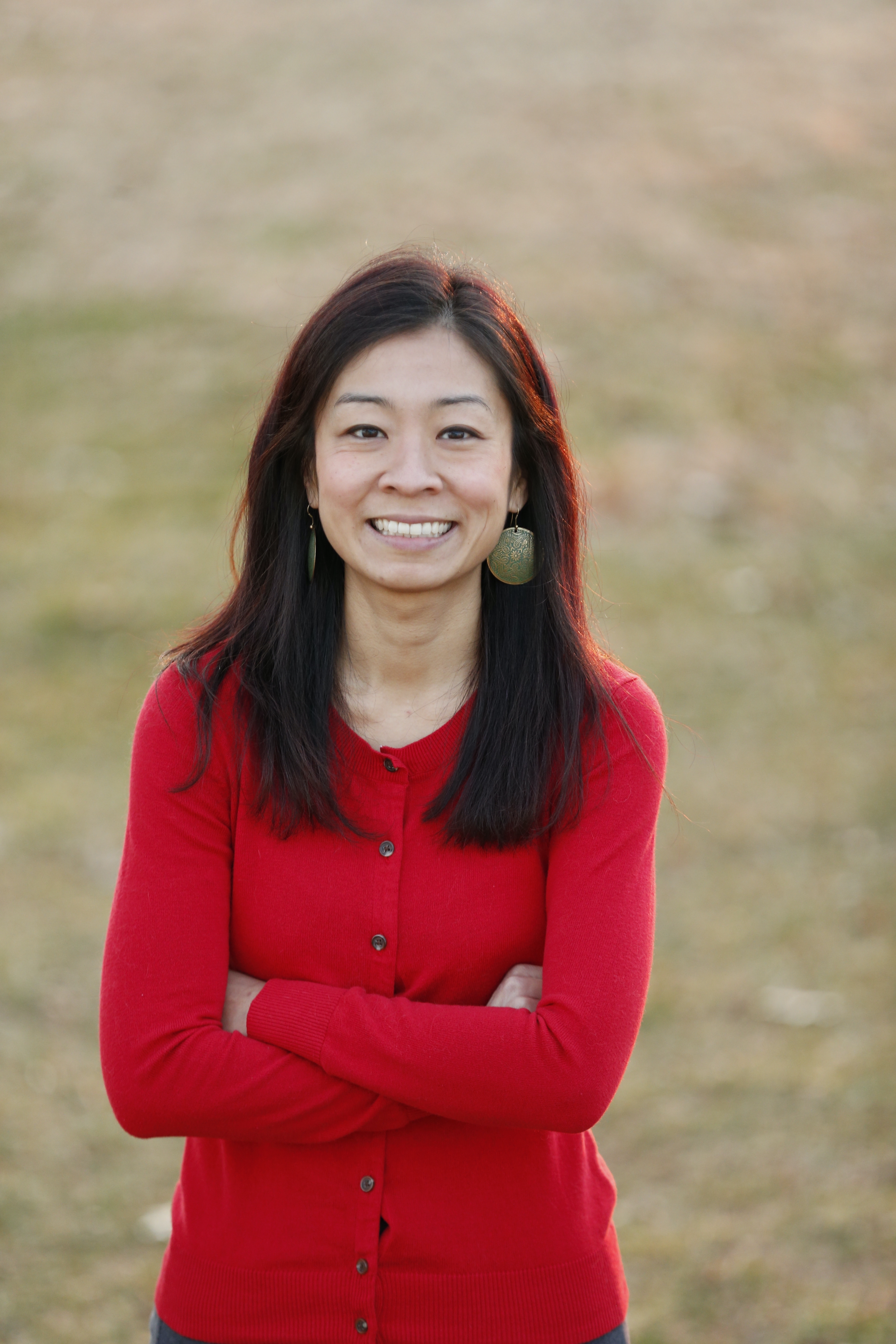 A young Asian woman smiles in a field of yellow and green grass, wearing a red cardigan with her arms folded.